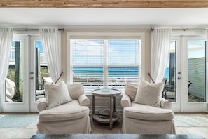 Ein Zimmer mit zwei beigen Sesseln, einem runden Tisch und Meerblick durch große Fenster mit Jalousien.