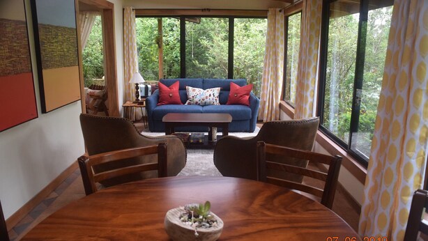 Una sala de estar acogedora con un sofá azul, cojines rojos y una mesa de centro. Se observan dos sillas de madera y una planta pequeña sobre la mesa.
