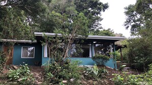 Una casa pequeña con porche, rodeada de vegetación y árboles frondosos.