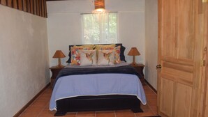 Un dormitorio con una cama, dos mesitas de noche, un ventanal con cortinas y una puerta de madera.