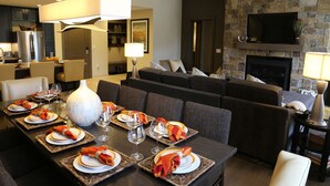 A dining area with a long table set for a meal, a sofa in the background, and a stone fireplace.