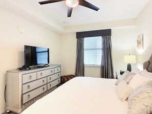 A hotel room with a bed, a dresser, a television, a ceiling fan, a window with curtains, and a bedside lamp. - Kissimmee Condo: Pools, parks, family fun, and attractions! (Kissimmee)