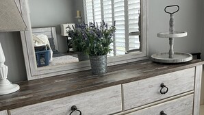 A wooden dresser with six drawers, each with a metal ring handle. A large mirror above the dresser reflects a room with a window and a plant.