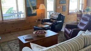 Ein gemütliches Wohnzimmer mit Holzwänden, einem gestreiften Sofa, Ledersesseln und einem großen Fenster mit Jalousien.