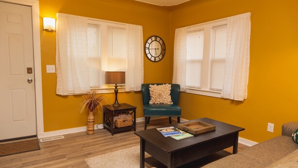 Un salon comprenant un canapé, une table basse et un fauteuil agrémenté d’un coussin décoratif. Il y a une horloge au mur et une lampe sur une table d’appoint.
