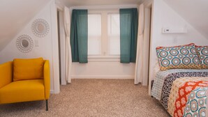 Une chambre avec une chaise jaune, un lit avec une literie à motifs, une fenêtre avec des rideaux verts et un miroir fixé au mur.