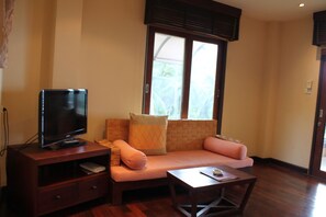 A living room with a television on a wooden stand, a sofa with cushions, a coffee table, and a window with blinds.