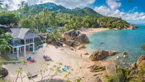 A beach with large rocks, palm trees, and a thatched-roof building with beach chairs and umbrellas. - Coral Cliff Beach Resort Samui (Koh Samui)