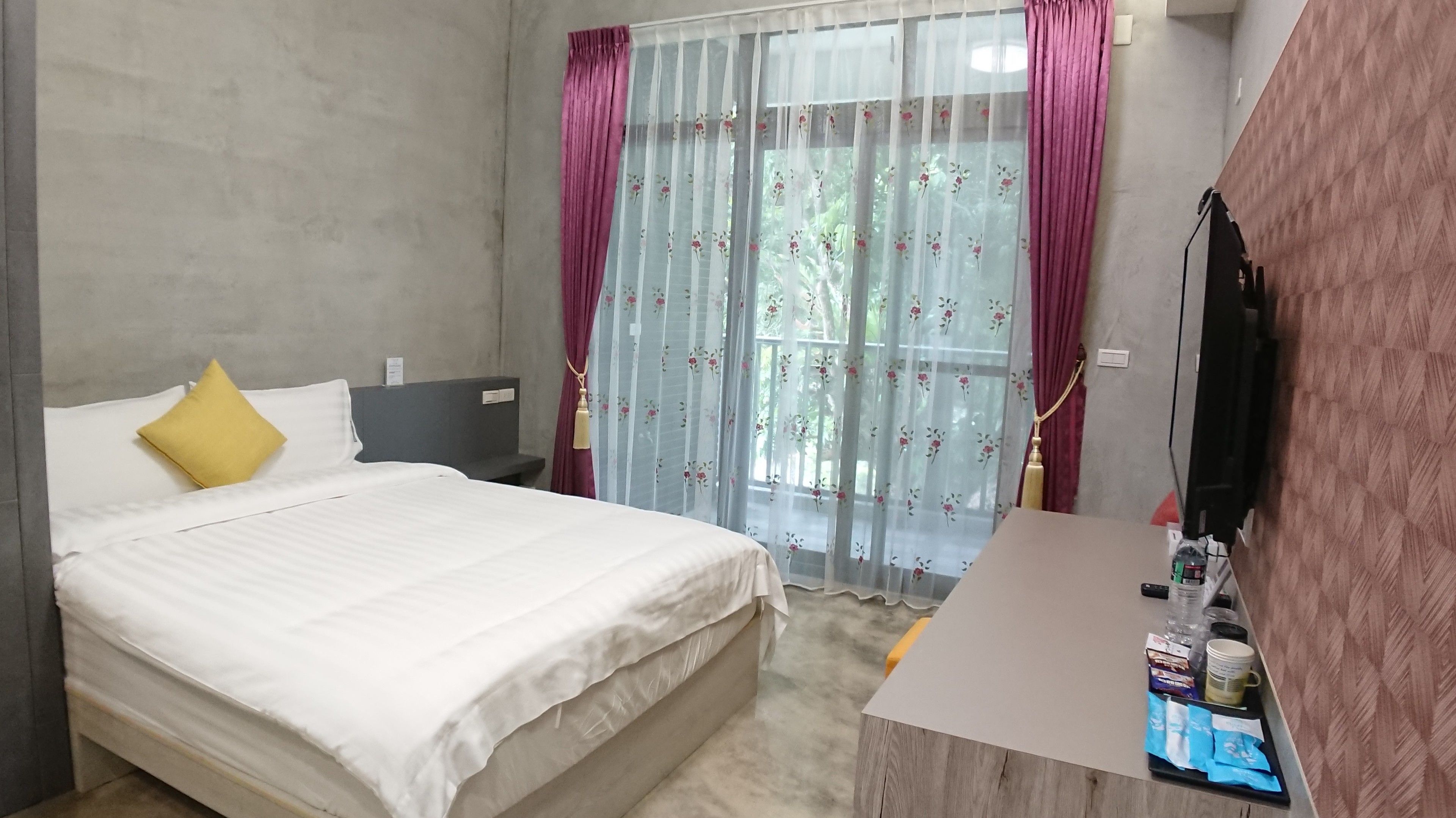 A hotel room with a bed, a bedside table with toiletries, a TV, and a window with sheer curtains.