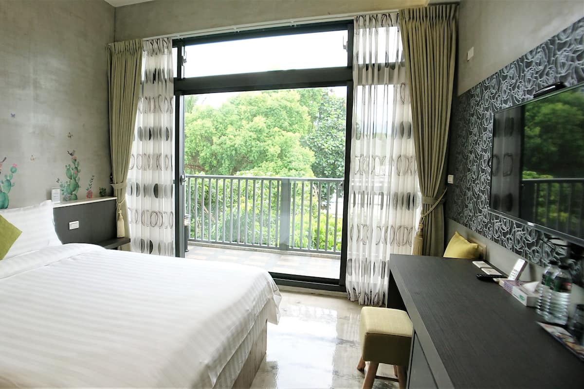 A hotel room with a bed, a desk, a chair, a large window with curtains, and a balcony with a railing.