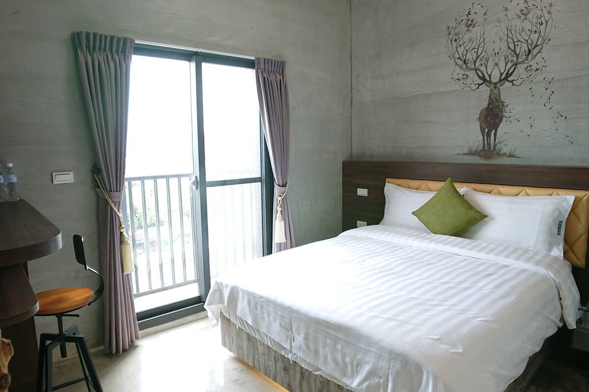 A hotel room with a bed, a desk, a chair, and a view of the outdoors through a sliding door.