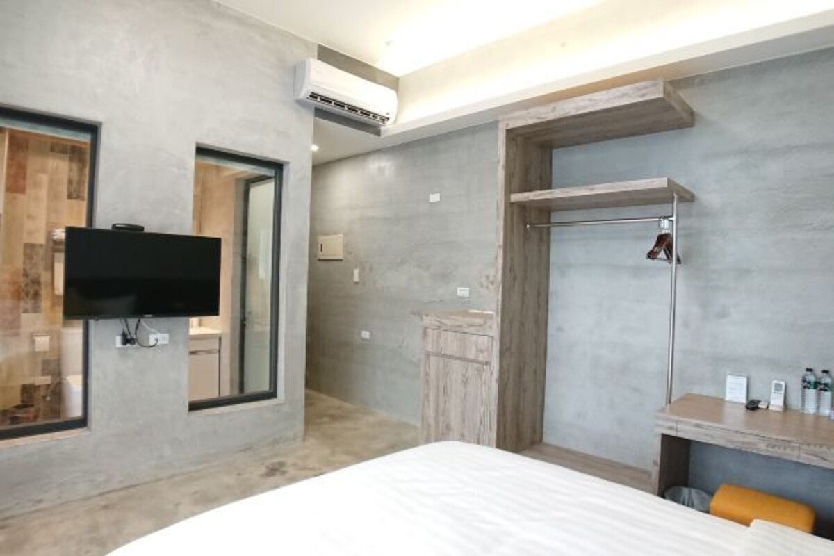 A modern hotel room with a large bed, a TV mounted on the wall, a wooden vanity, and a shower area.