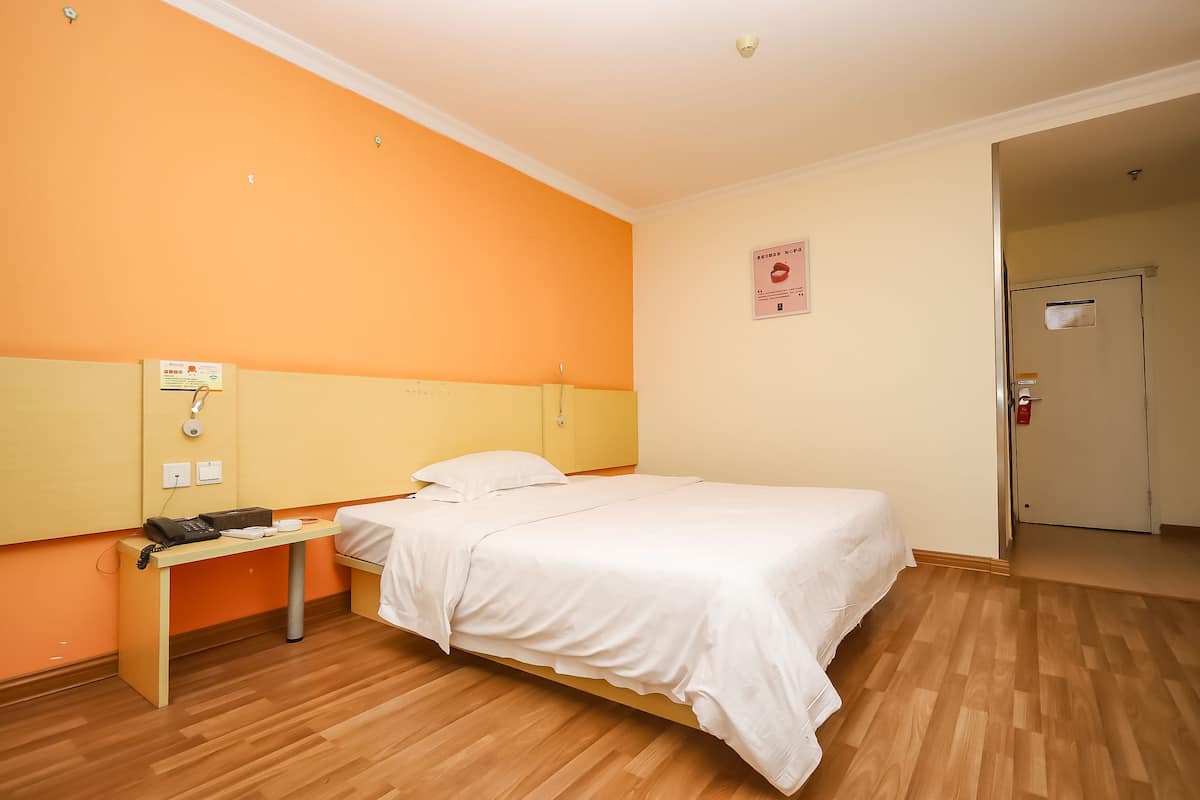 A hotel room with a single bed, a bedside table with a phone and a cup, an orange wall, and a door to the right.