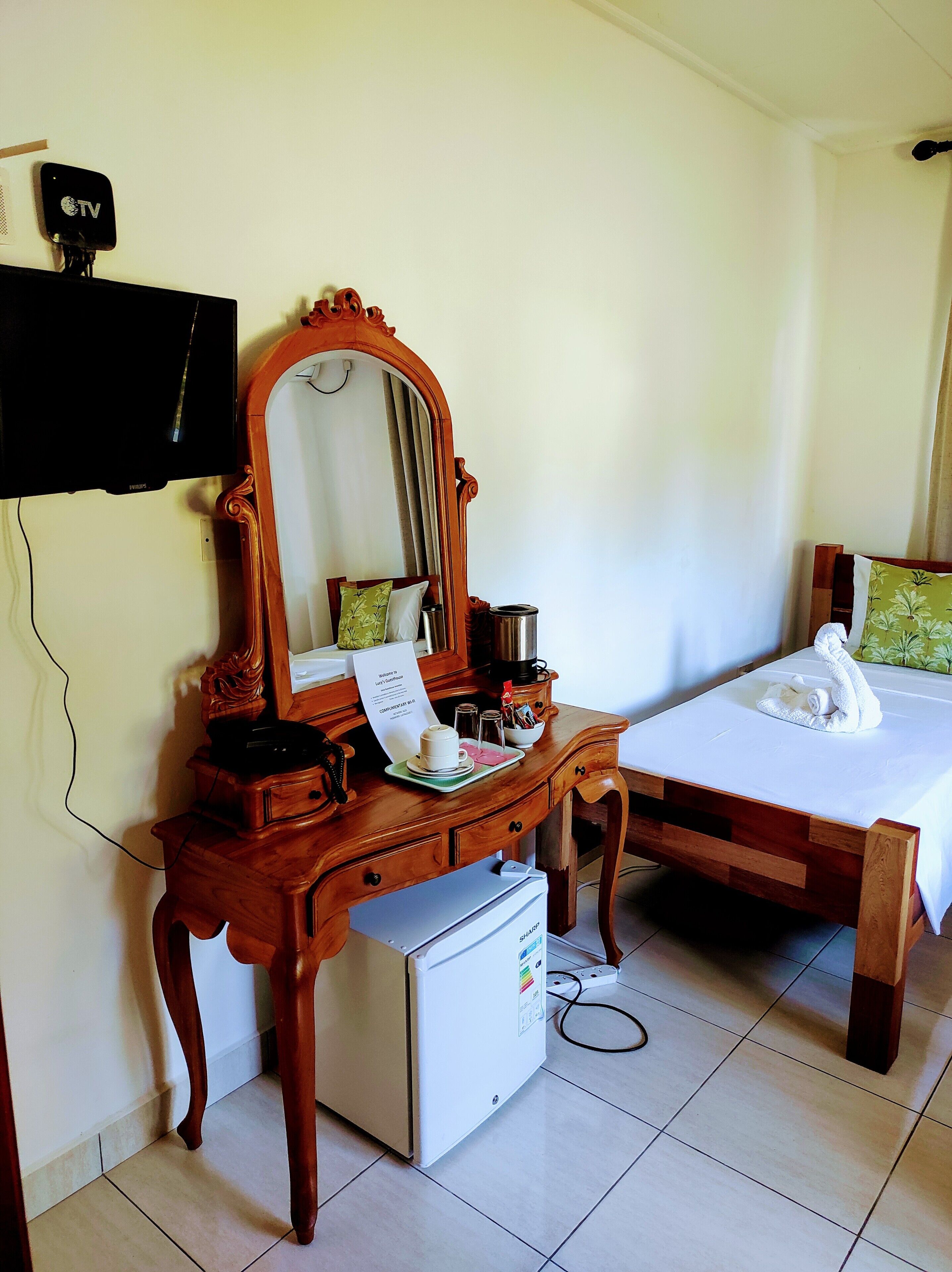 Habitación de hotel con cómoda de madera, espejo, refrigerador pequeño, cama con cubrecama blanco y televisor montado en la pared.