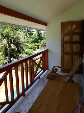 A balcony with a wooden railing, a wooden table, and a chair. The balcony overlooks a lush garden with palm trees and a view of the sky. - Lucy's guesthouse (La Digue)
