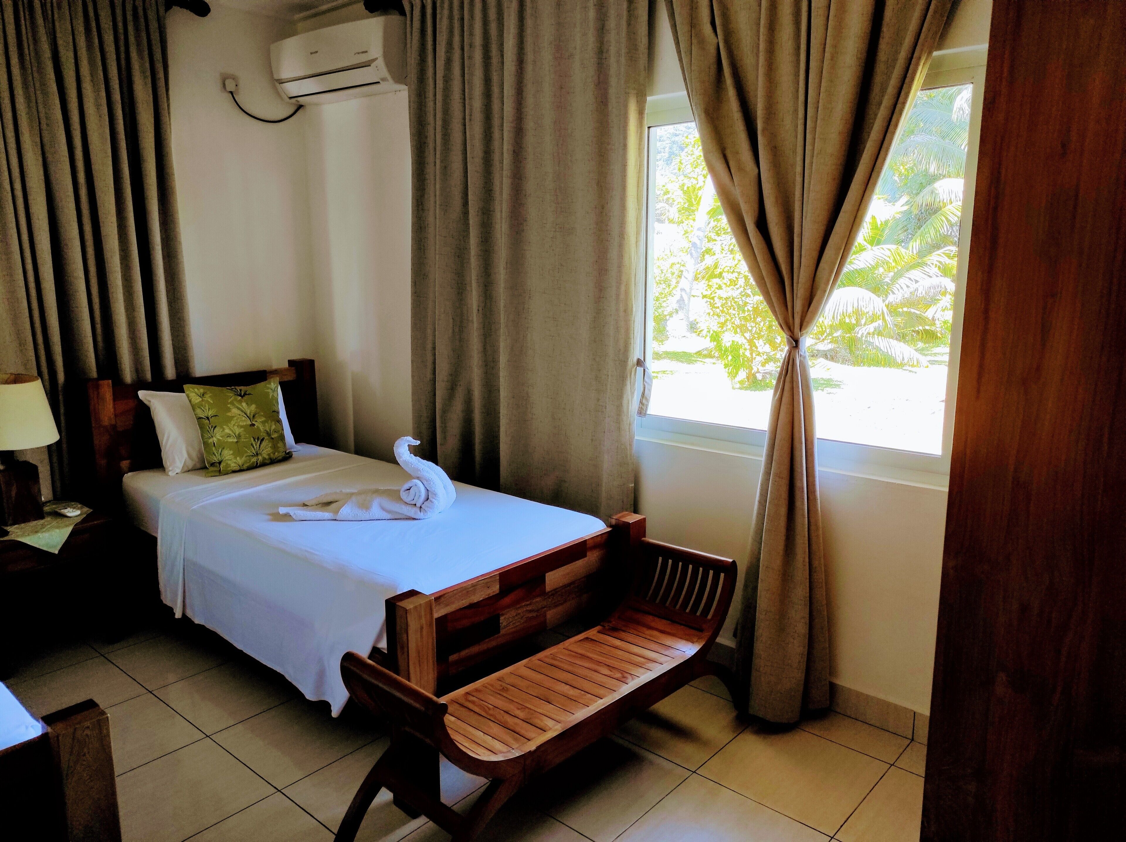A hotel room with a bed, a wooden bench, a nightstand, a lamp, and a window with curtains.