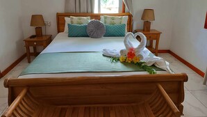 A neatly made bed with a wooden headboard, two bedside tables with lamps, and a window with curtains. - Lucy's guesthouse (La Digue)