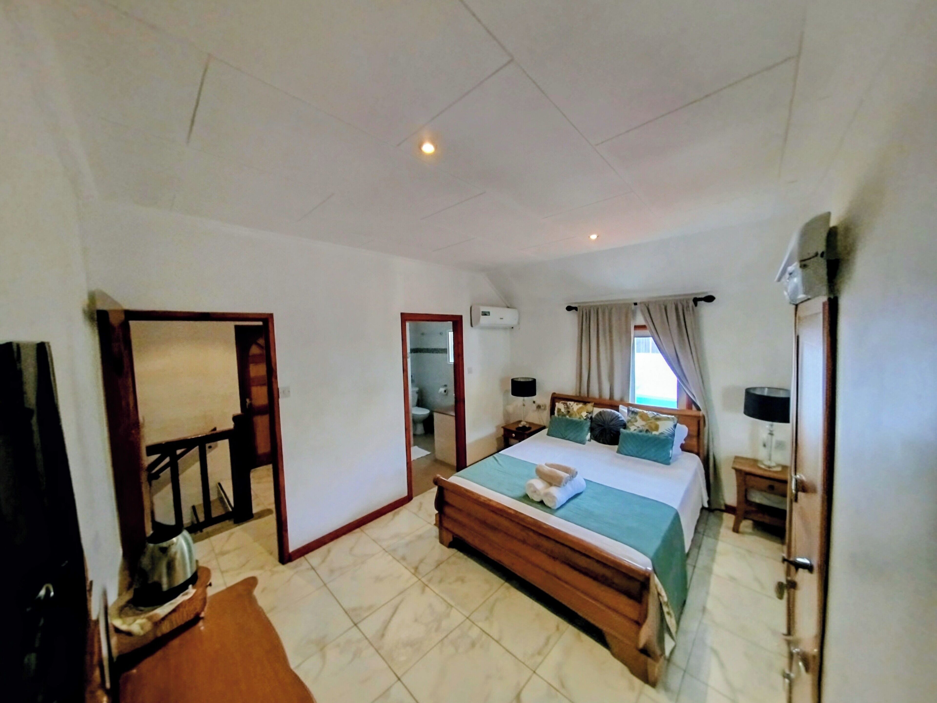 A spacious bedroom with a large bed, a wooden headboard, a bedside table with a lamp, and a view of the outside through a window with curtains.