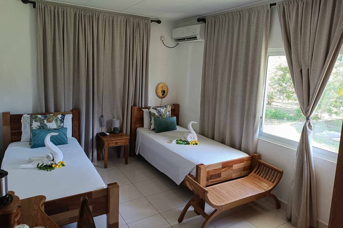 A hotel room with two beds, a wooden chair, a small table, and a window with curtains.