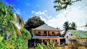 A two-story house with a balcony, surrounded by lush greenery and a clear sky. - Lucy's guesthouse (La Digue)