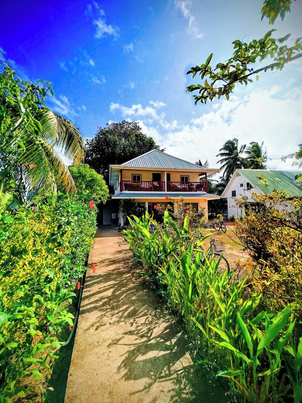 A two-story house with a balcony, surrounded by lush greenery and a clear sky. - Lucy's guesthouse (La Digue)
