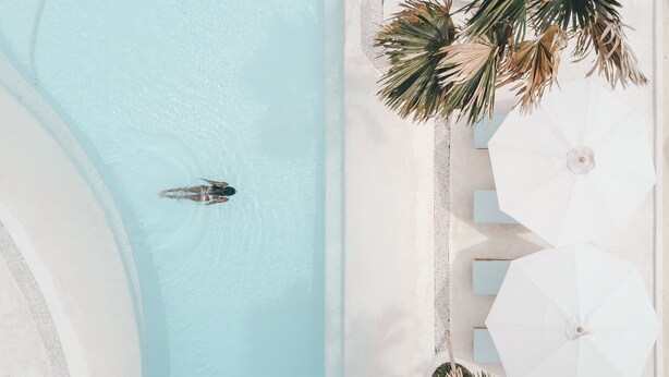 Vista aérea de una piscina con una persona nadando, rodeada de palmeras y sombrillas blancas.