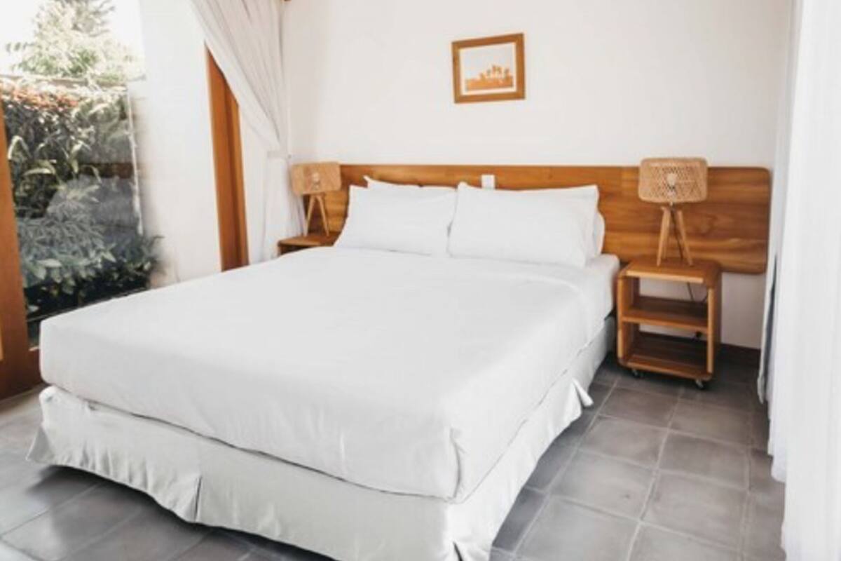 A bedroom with a large bed, wooden headboard, two bedside tables with lamps, a picture on the wall, and a view of greenery outside.