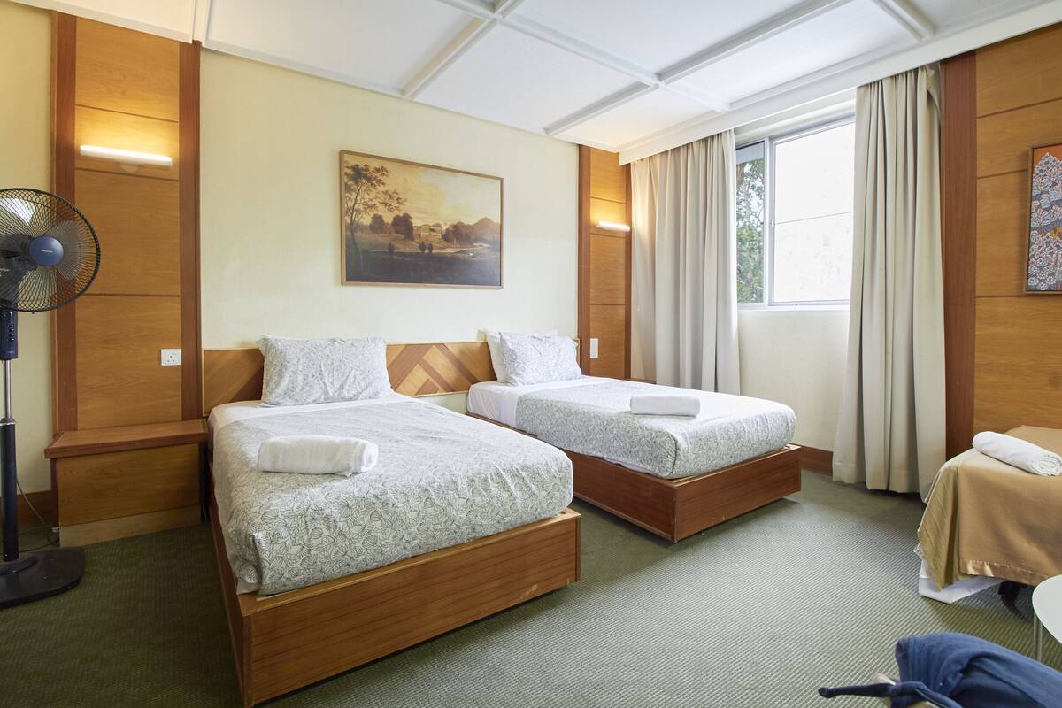 A hotel room with two wooden beds, a standing fan, a painting on the wall, and a window with curtains.