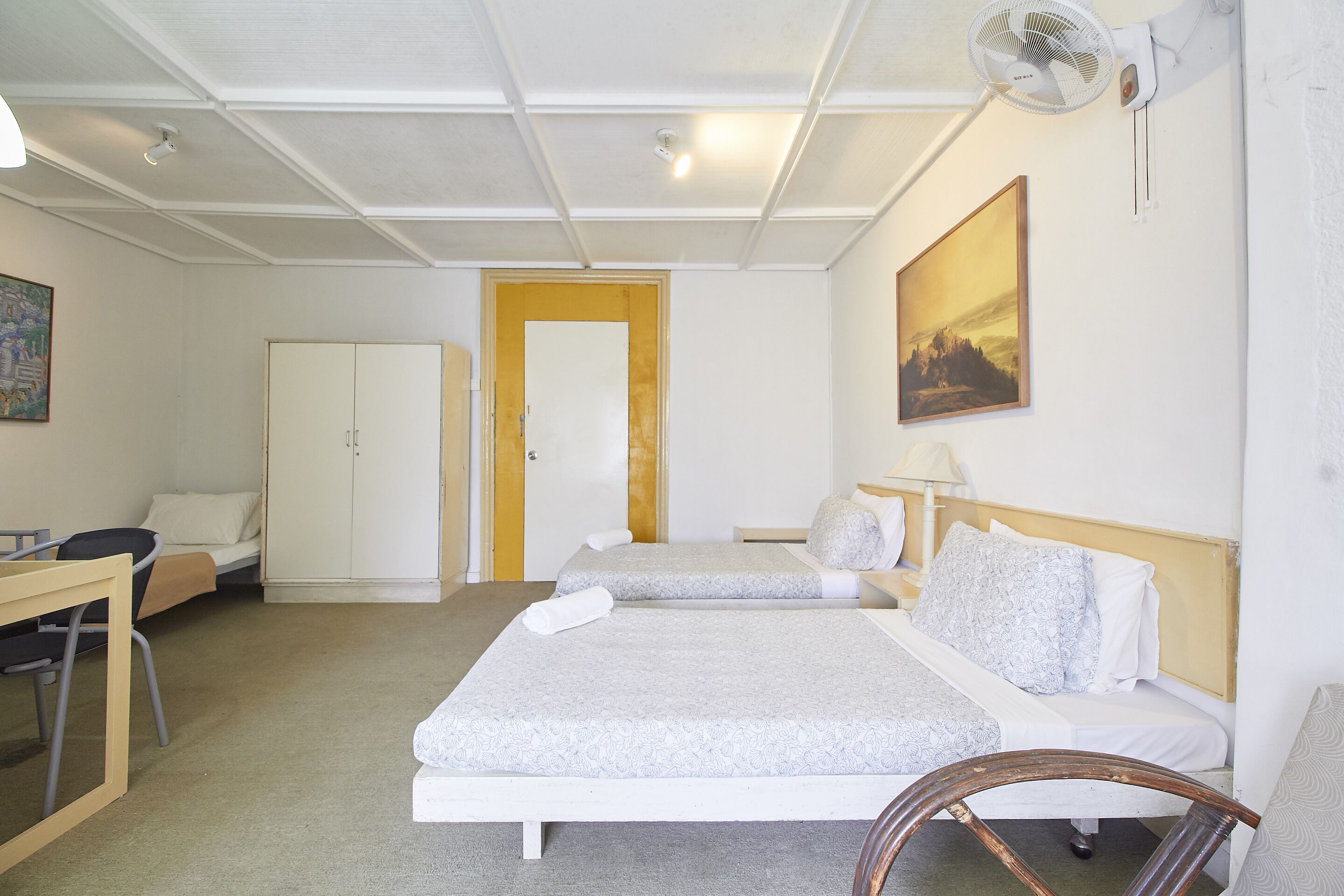 Habitación de hotel con dos camas, un escritorio, una silla, un armario y un ventilador de techo.