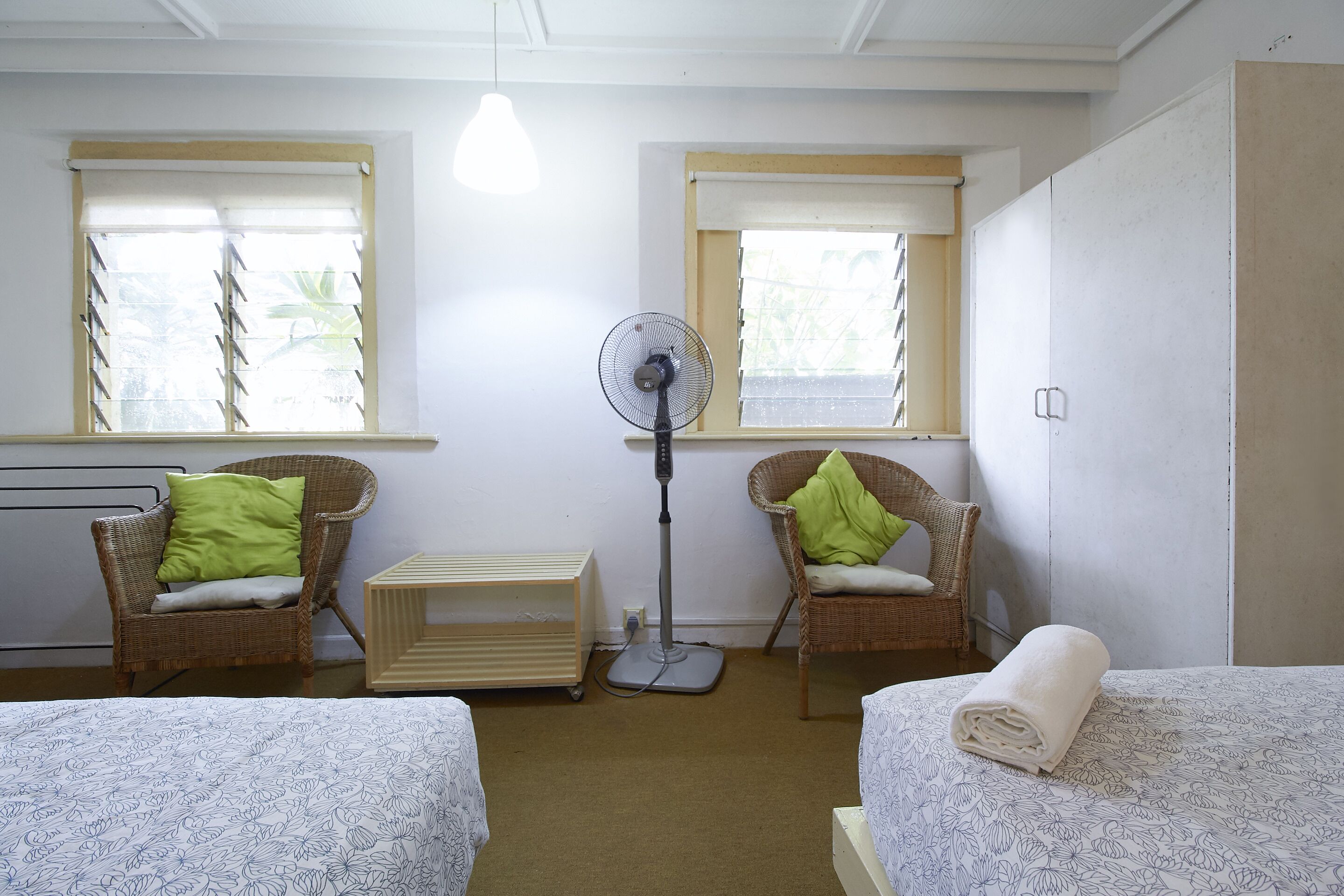 A room with two single beds, a standing fan, two wicker chairs with cushions, a radiator, and a window with blinds.