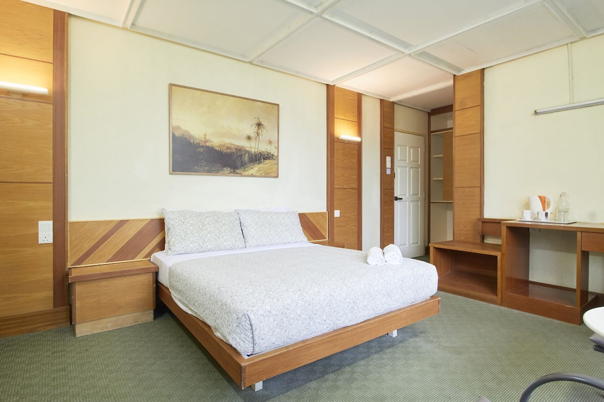 A hotel room with a wooden bed, a painting on the wall, a desk, and a chair.