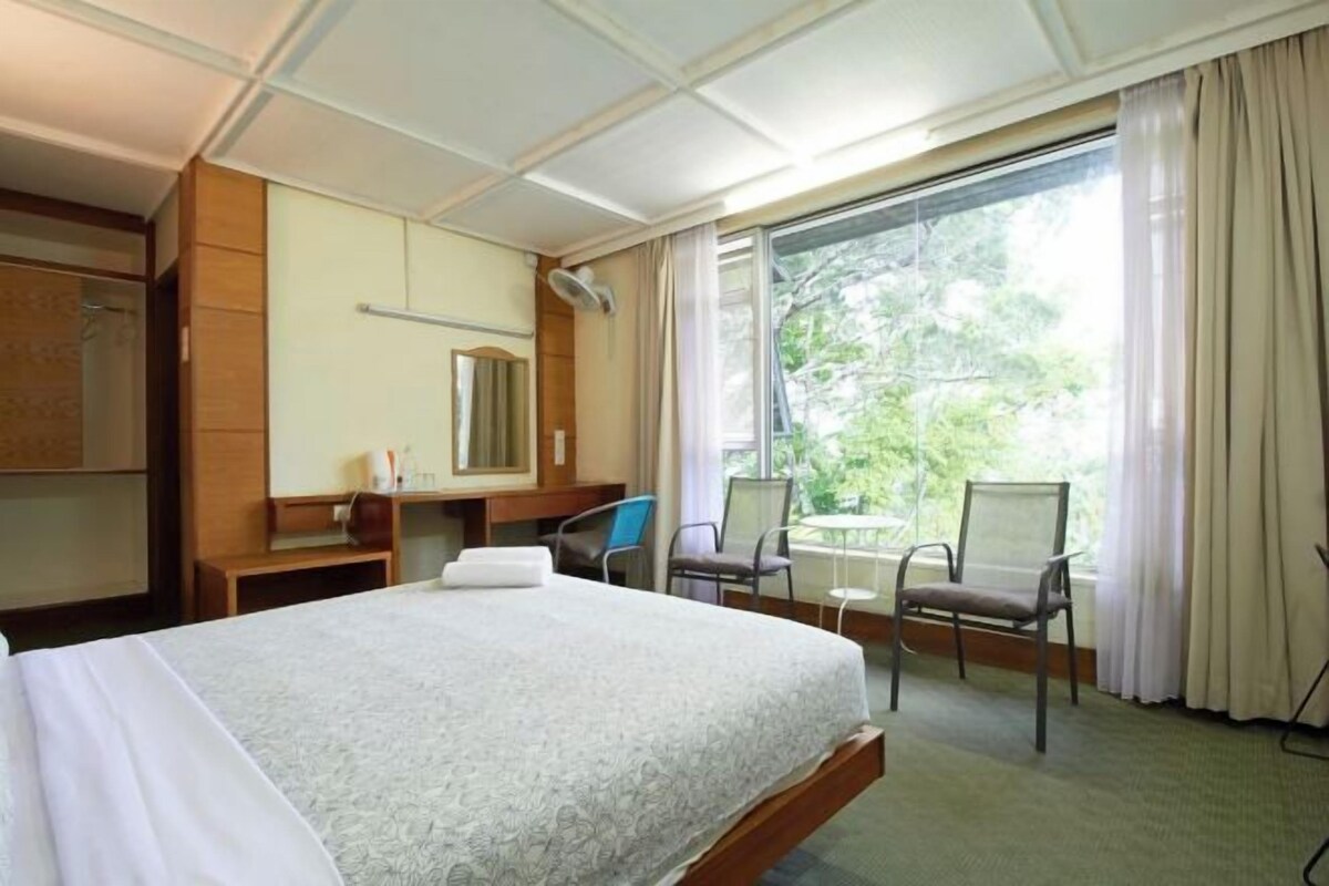 Una habitación de hotel con cama, escritorio, silla, mesita, espejo y ventana con cortinas.
