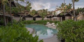 Ein Swimmingpool, umgeben von üppiger Vegetation und Bungalows auf Stelzen mit Außenbereich.