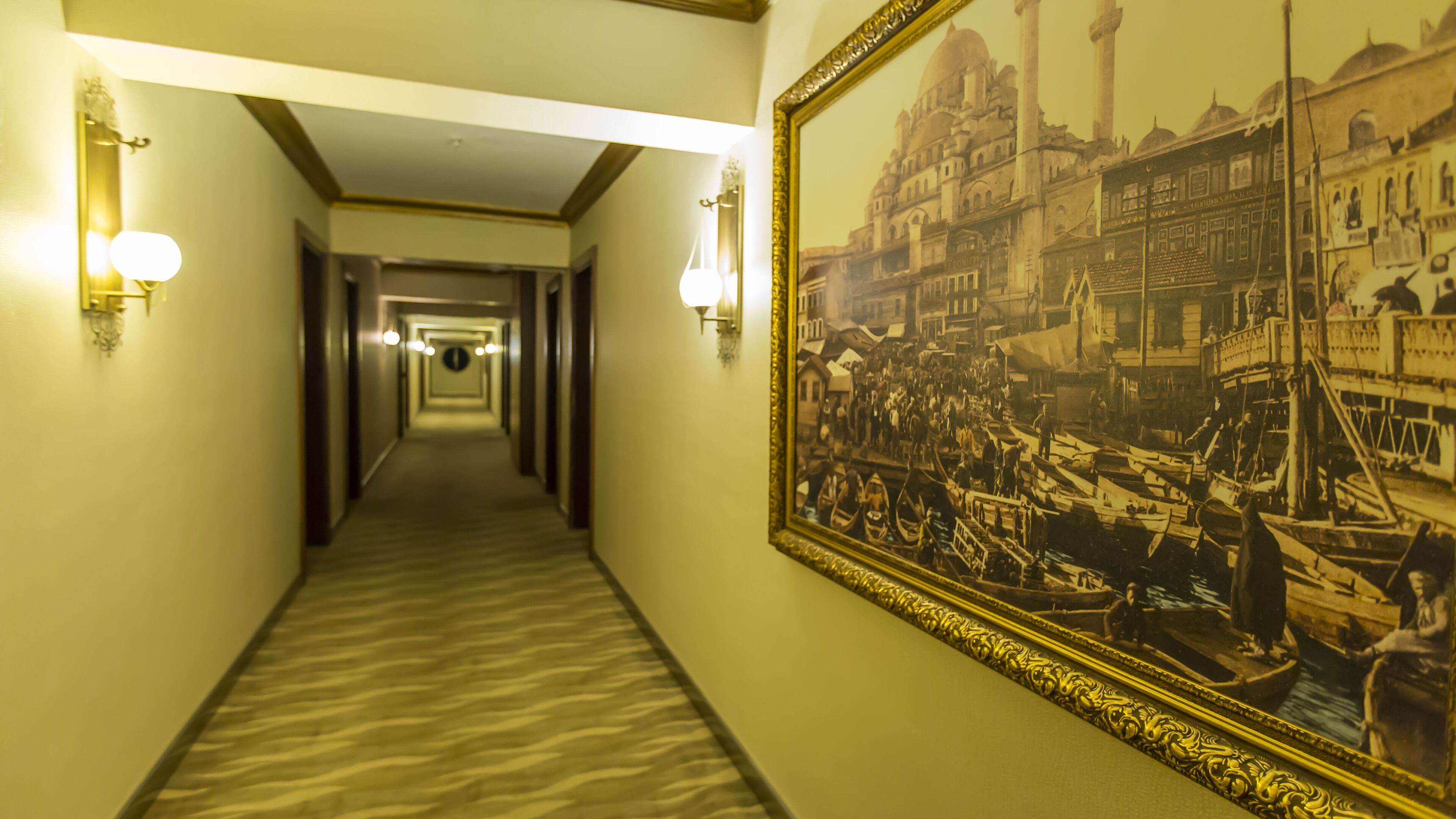 Un pasillo de hotel con piso alfombrado, luces fijadas a la pared y un cuadro histórico grande enmarcado a la derecha.