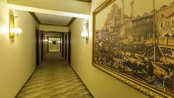 Un pasillo de hotel con piso alfombrado, luces fijadas a la pared y un cuadro histórico grande enmarcado a la derecha.