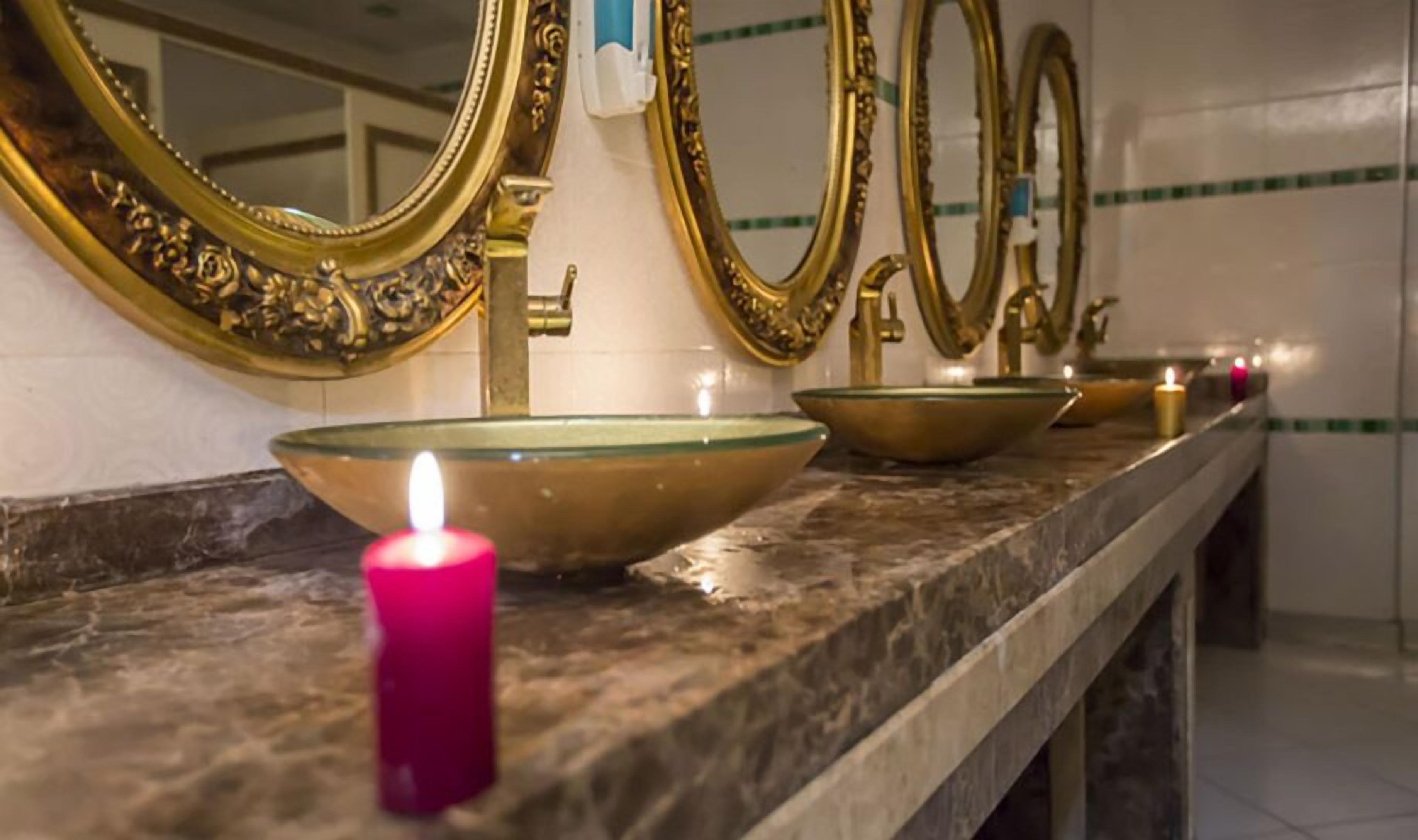 A bathroom with three gold-framed mirrors, a pink candle, and two glass bowls on a marble countertop.