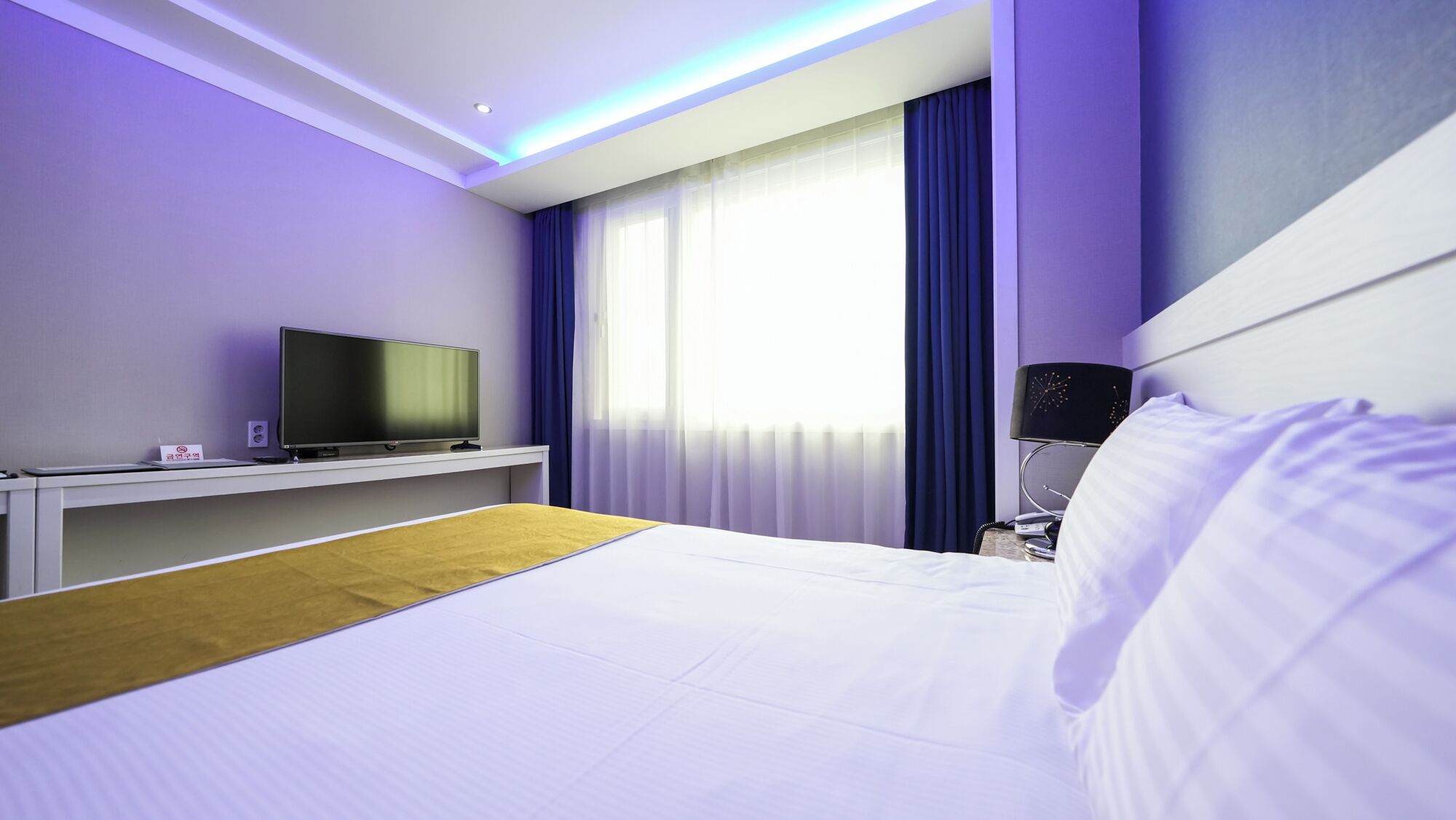Quarto de hotel moderno com uma cama, uma televisão sobre um móvel, uma mesa de cabeceira e uma janela com cortinas.