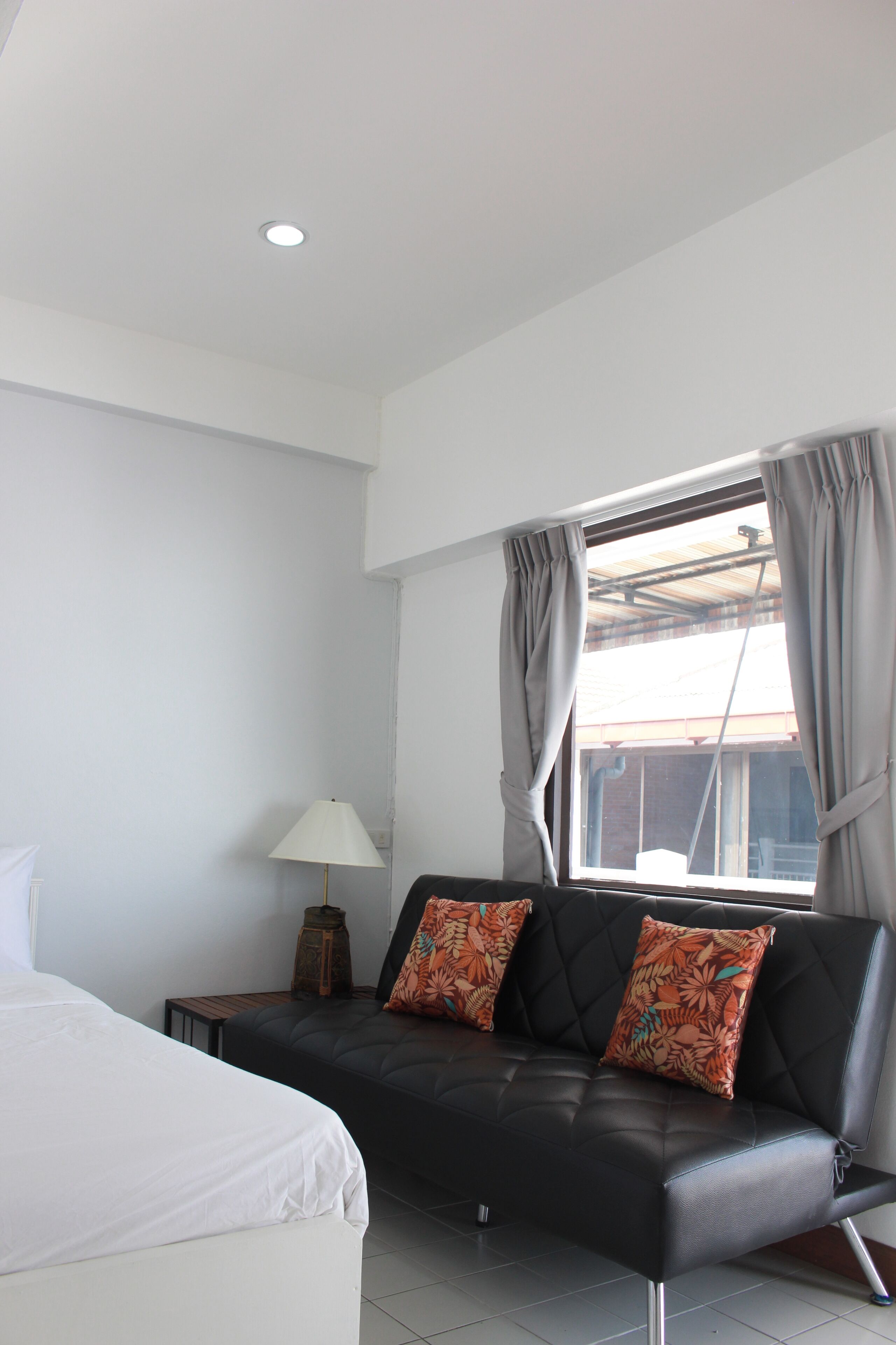 A modern hotel room with a bed, a black leather sofa with floral cushions, a small table with a lamp, and a window with curtains.