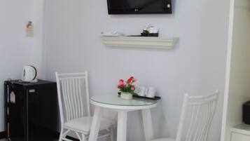 Een kleine, schone kamer met witte wanden, een flatscreen televisie bevestigd boven een witte plank, een klein rond tafeltje met twee witte stoelen en een zwarte koelkast.