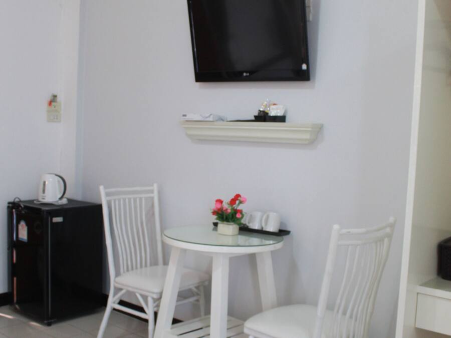 Een kleine, schone kamer met witte wanden, een flatscreen televisie bevestigd boven een witte plank, een klein rond tafeltje met twee witte stoelen en een zwarte koelkast.