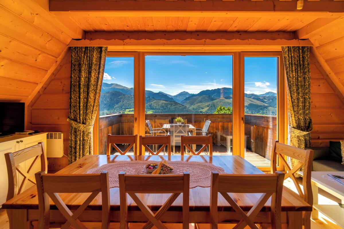 A wooden cabin with a dining area featuring a table and chairs, large windows with curtains, and a view of mountains.
