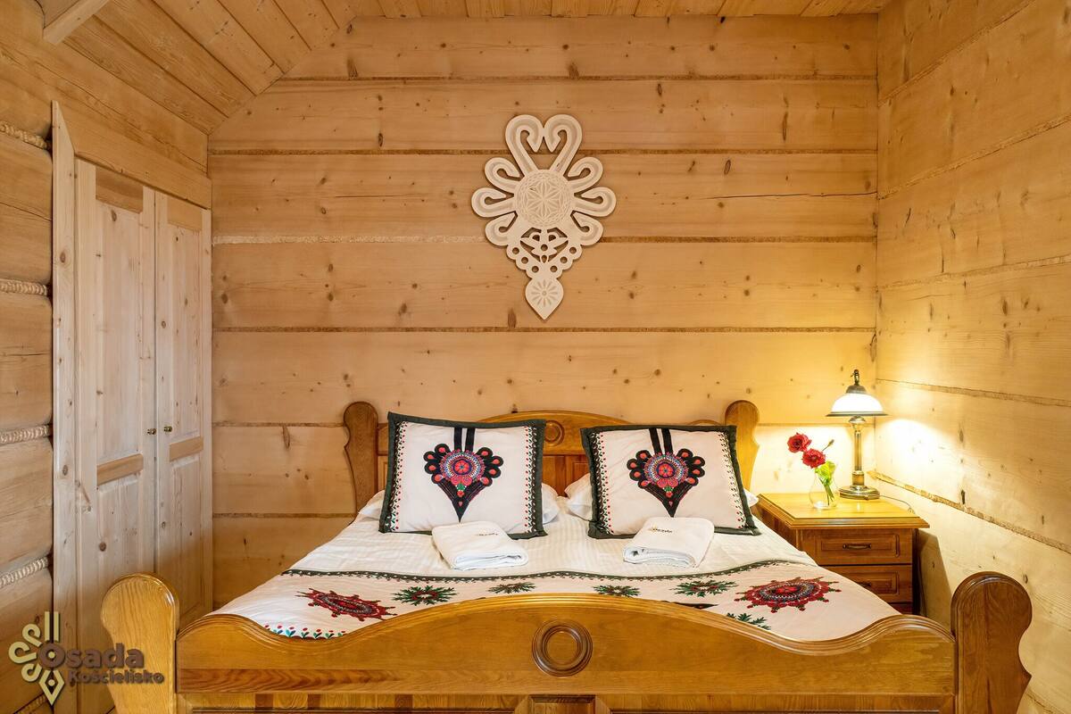 A wooden cabin room with a bed, two pillows, a nightstand with a lamp, and a decorative wall piece.