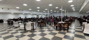 Um salão amplo com fileiras de mesas e cadeiras, piso xadrez e teto alto com iluminação.