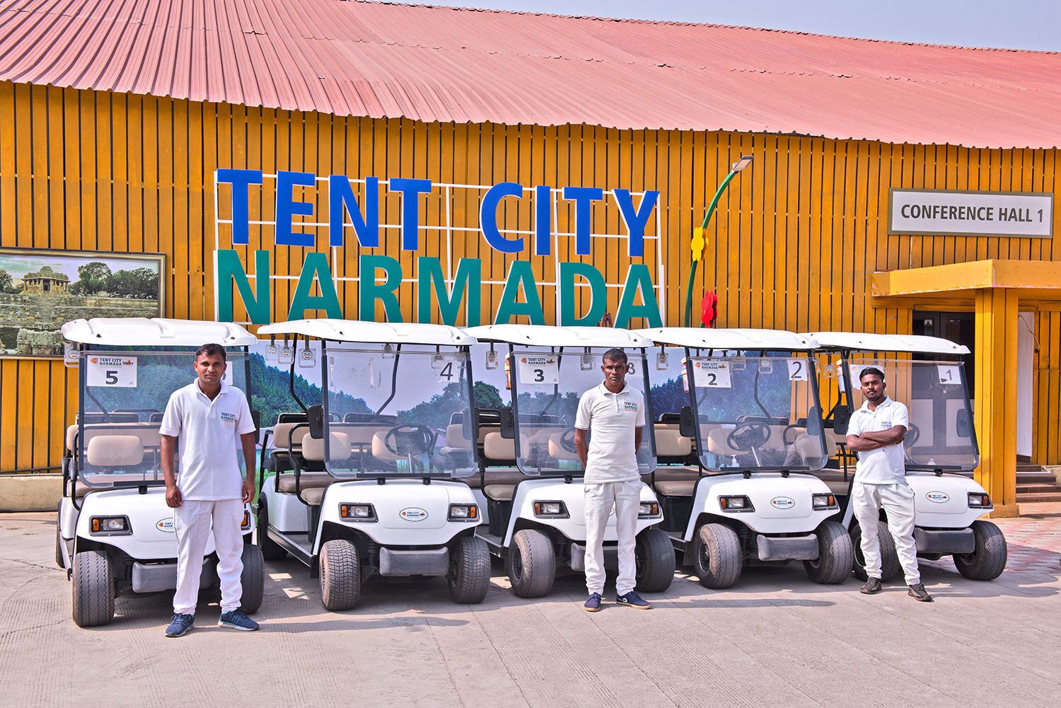 Flere golfbiler foran en bygning med et skilt, der står 'TENT CITY NARMADA'.