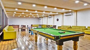 Una mesa de billar en una sala con sofás amarillos, suelo de madera y luces empotradas en el techo.