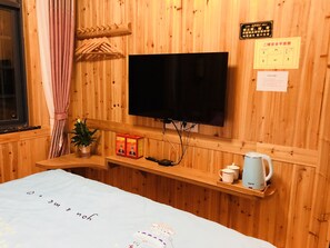 A wooden room with a bed, a TV mounted on the wall, a shelf with a remote control, a plant, and a kettle.