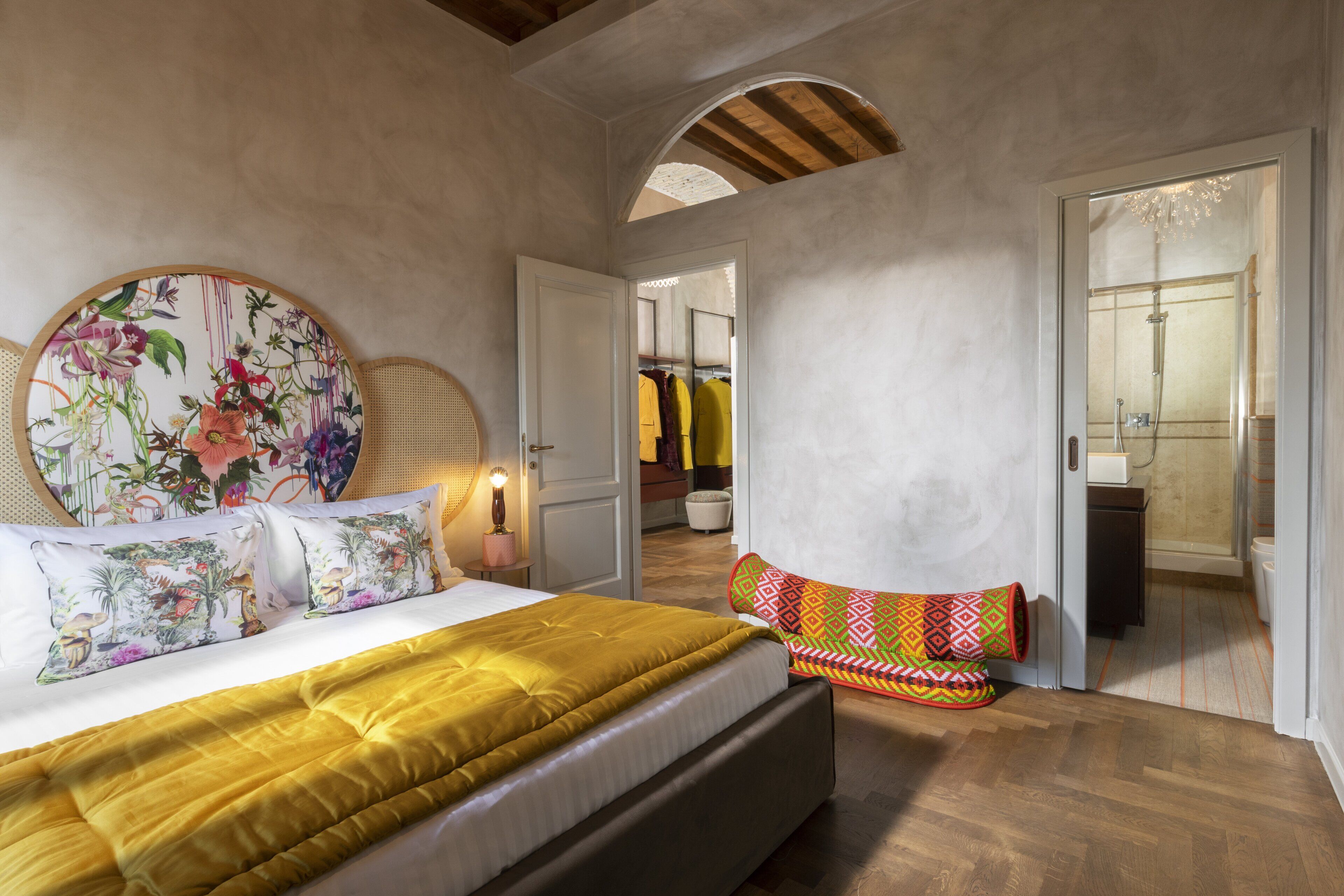 A bedroom with a large bed, a colorful headboard, a bench with a patterned cushion, and a walk-in closet with hanging clothes.