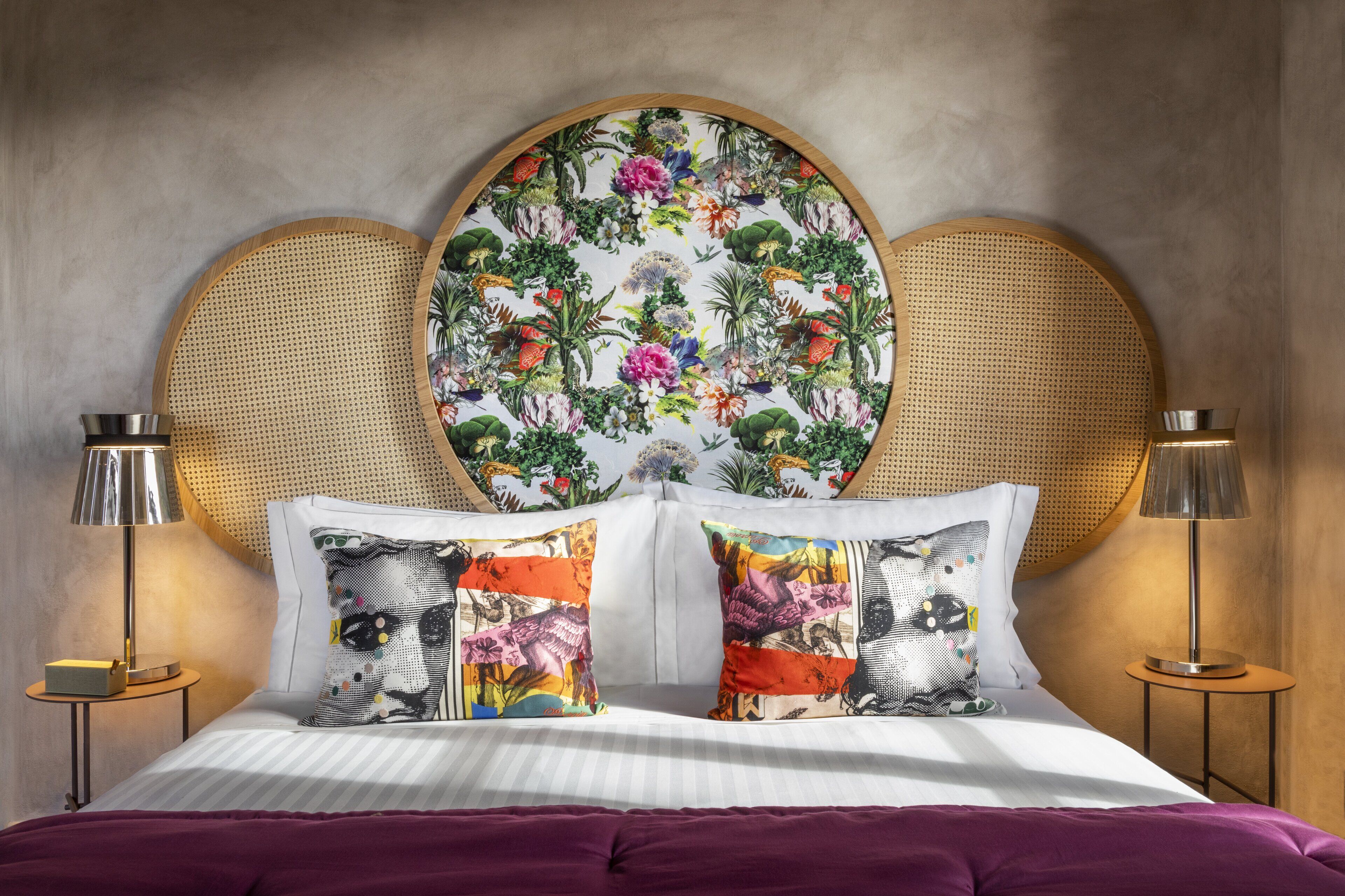 Una cama con cabecera estampada con motivos florales, dos lámparas de noche y una manta morada.