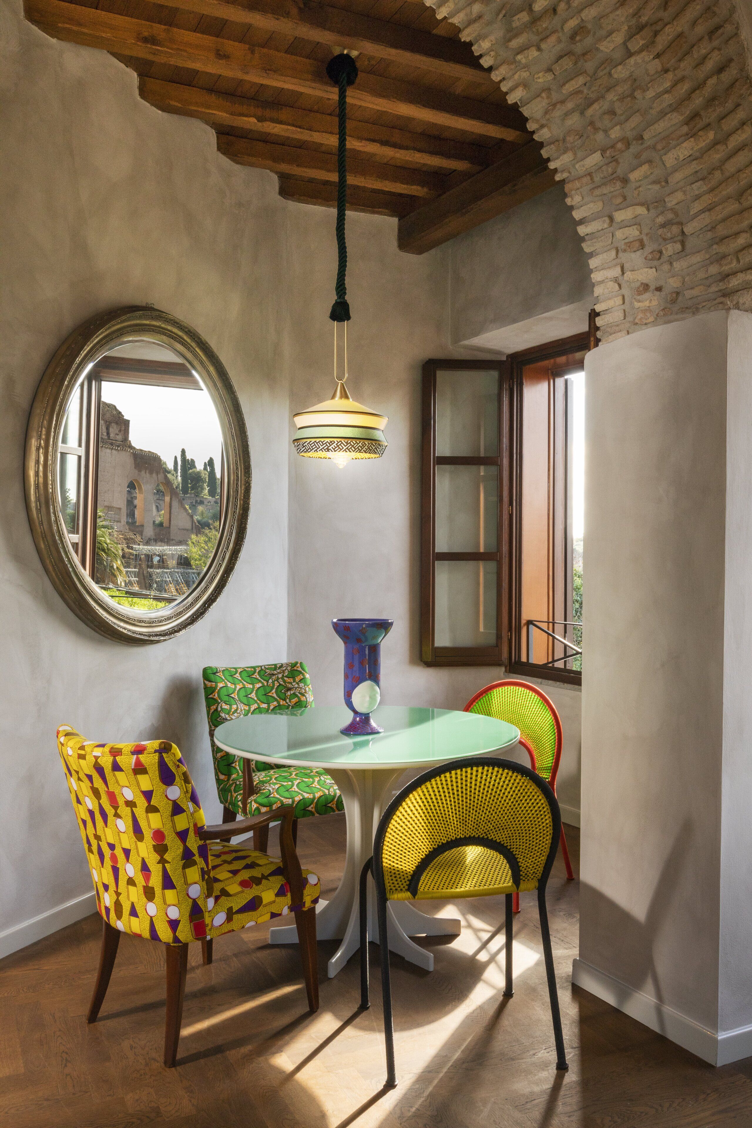 A dining area with a round table, colorful chairs, a hanging light fixture, and a view of an old building through a window.