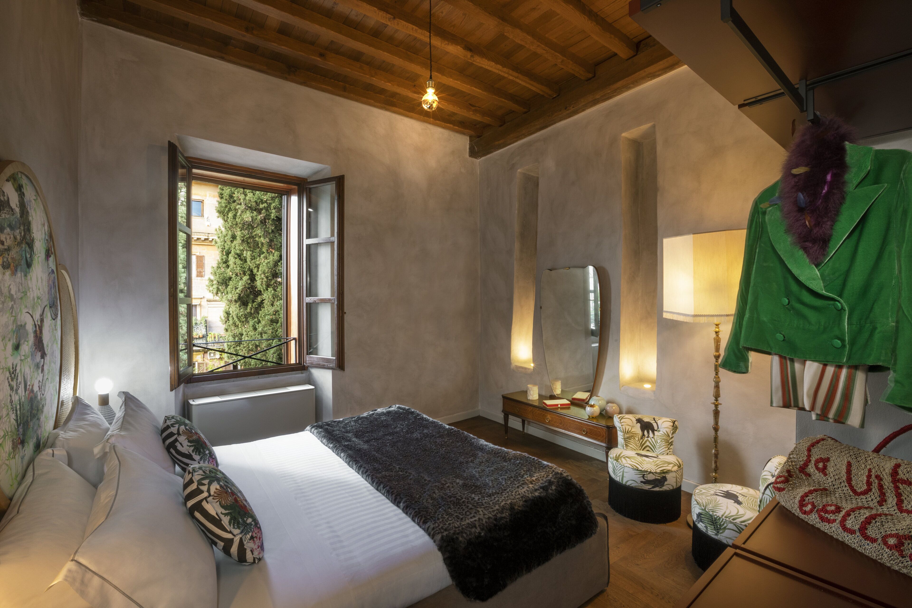 Ein Schlafzimmer mit Bett, Fenster, Spiegel, Lampe und Stuhl. Ein grüner Mantel hängt an der Wand.